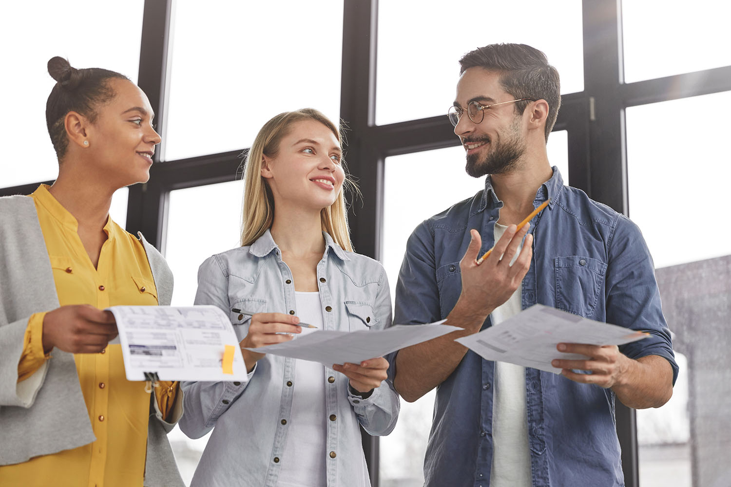 Group of friendly three multiethnic business people stand in office hall, hold papers, study financial report, try come to solution how clear debts, have pleased expressions as going to have corporate